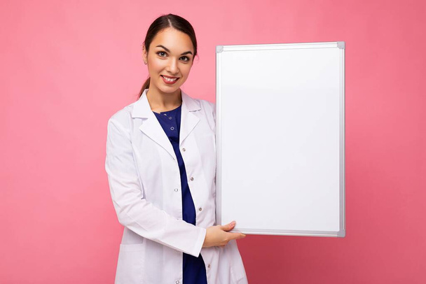 jong van mooie gelukkig brunet vrouw dragen medische witte jas met witte magneetbord voor mockup geïsoleerd op roze achtergrond met lege ruimte - Foto, afbeelding