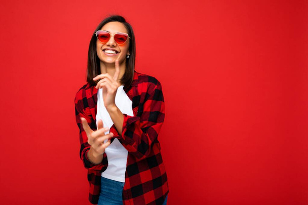 Photo of young beautiful happy smiling brunette woman wearing trendy white t-shirt and red check shirt and red sunglasses. Sexy carefree female person posing isolated near red wall in studio with free - Photo, Image