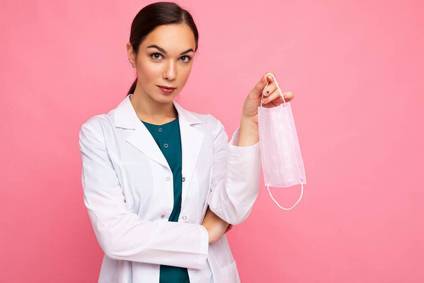 Portret van een aantrekkelijke jonge vrouwelijke arts in witte jas met medisch masker geïsoleerd op roze achtergrond - Foto, afbeelding