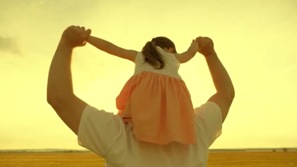 Padre cammina con la figlia sulle spalle in raggi di tramonto. La famiglia felice sta riposando nel parco. Papà porta il suo amato bambino al collo al sole. Un bambino con un genitore cammina nel fine settimana - Filmati, video