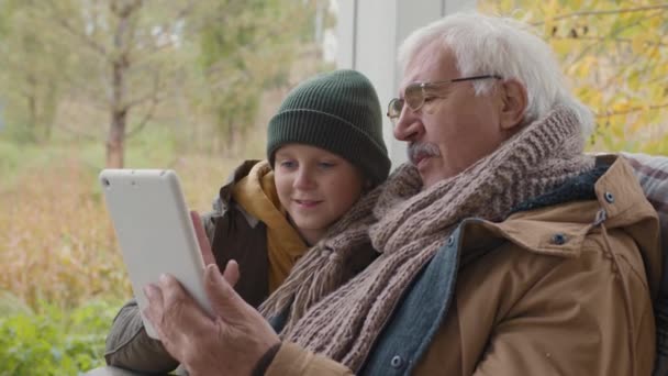 歳の白人男性と彼の十代の孫のロックダウン両方の暖かい服を着てテラスに座って、デジタルタブレットを使用してビデオ会話をして - 映像、動画