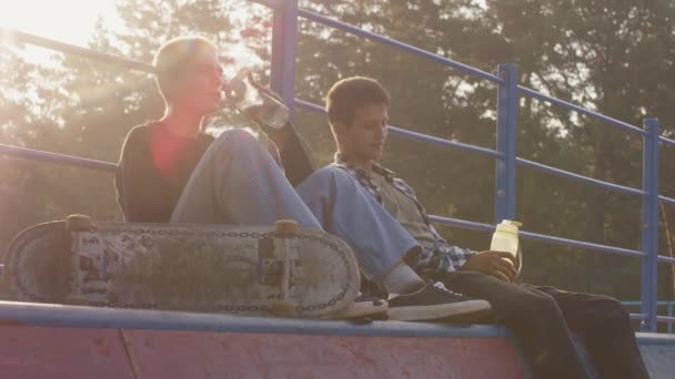 Kézi lassított felvétel tinédzserekről, akik a rámpa tetején ülnek a skateparkban, és korcsolyázás után isznak sportvizes palackokból. - Felvétel, videó