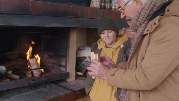 Μέτριο πλάνο του ηλικιωμένου Καυκάσου άνδρα που φοράει ζεστό σακάκι και μαντήλι κρατώντας μικρά κομμάτια ξύλου και μαθαίνοντας στον εγγονό του πώς να ανάβει ένα υπαίθριο τζάκι - Πλάνα, βίντεο