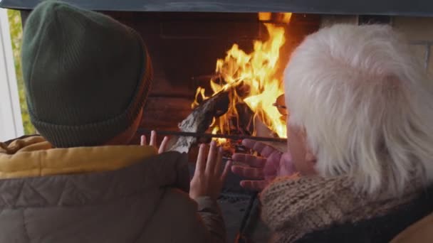 Back view of teenage Caucasian boy wearing warm clothes and his grandfather wearing eyeglasses standing at outdoor fireplace, warming hands by fire and having talk - Footage, Video