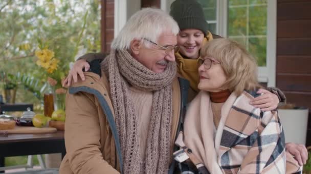 Medium shot van gelukkige oude blanke echtgenoten in warme kleren zittend op het terras van hun huis dan komt hun kleinzoon, knuffelend hen en iedereen begint te lachen - Video