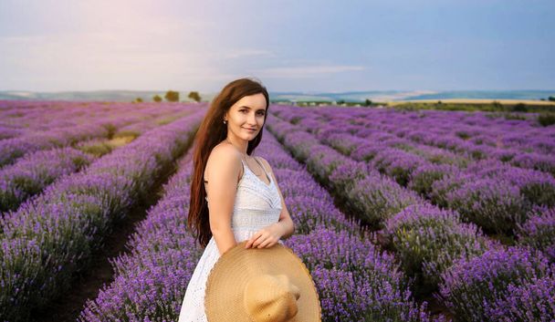 Portrait of a young woman in white dress and straw hat walking in the lavender field. Lavender flowers in bloom. - Photo, Image