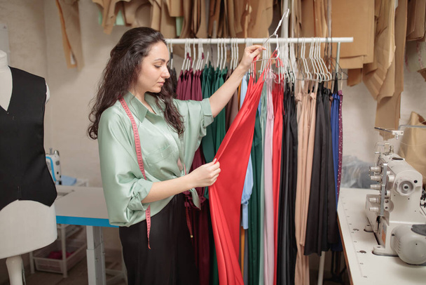 Female fashion designer works on new womenswear collection for clients in cozy workshop studio, dressmaker, tailor or needlewoman standing near clothing rack with fashionable stylish handmade clothes. Small business cocept - Photo, Image