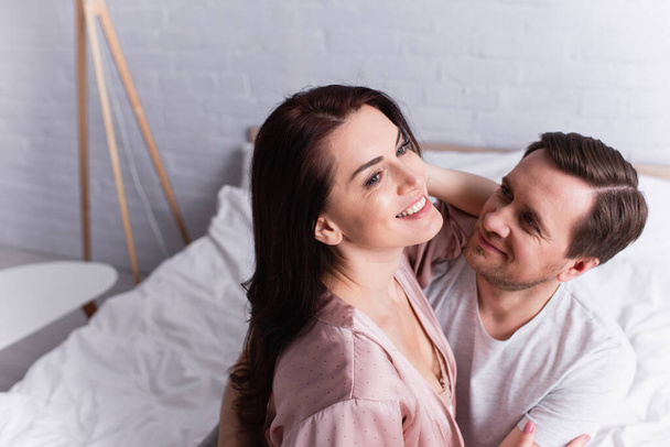 Smiling man hugging wife on bed on blurred background  - Valokuva, kuva