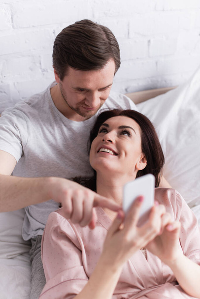Vista di angolo alto di uomo che punta a smartphone in mani di moglie allegra  - Foto, immagini