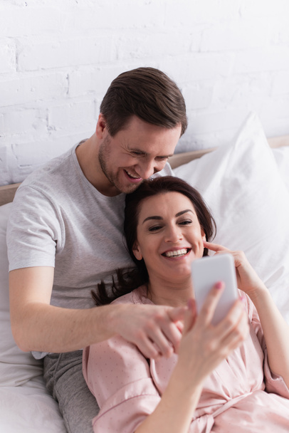 Smiling adult couple using smartphone on blurred foreground on bed  - Фото, изображение