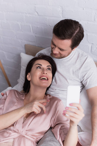 Smiling woman pointing at smartphone on blurred foreground near husband on bed  - Fotografie, Obrázek