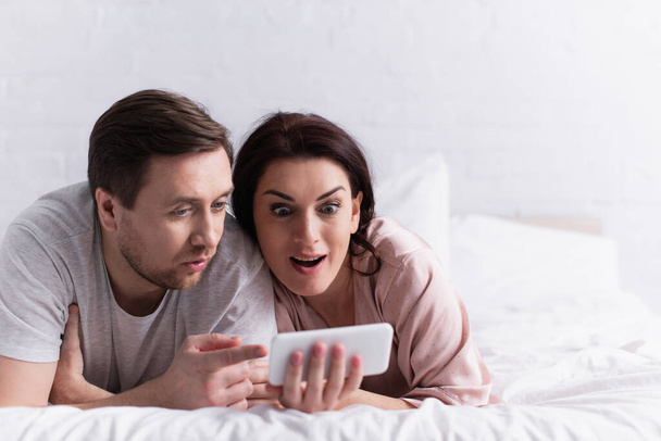 Couple adulte excité regardant smartphone au premier plan flou sur le lit  - Photo, image