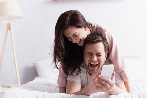 Laughing man holding cellphone on blurred foreground near wife on bed in morning  - Foto, Bild