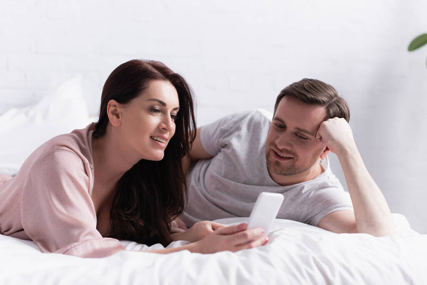 Brünette Frau hält Smartphone neben Ehemann im Schlafzimmer  - Foto, Bild
