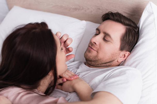 Homme adulte touchant le visage de la femme brune au premier plan flou sur le lit  - Photo, image