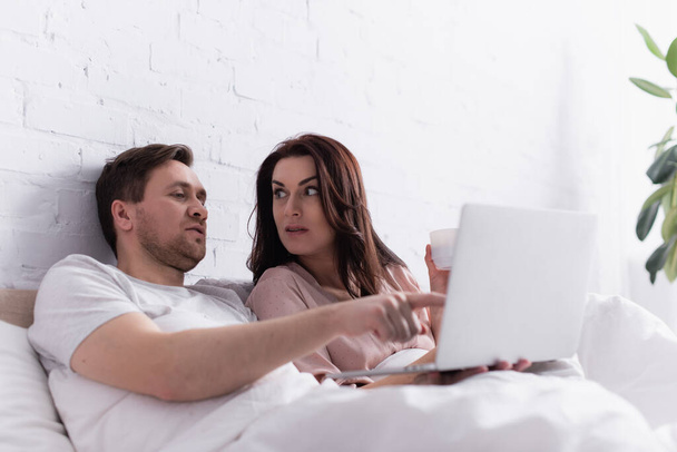 Man pointing with finger at laptop near wife with cup on bed  - 写真・画像