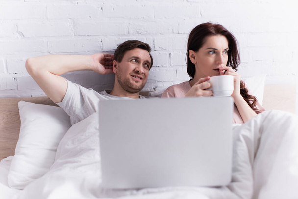 Stresszes nő kezében csésze kávét közel férj és laptop elmosódott előtérben az ágyon  - Fotó, kép