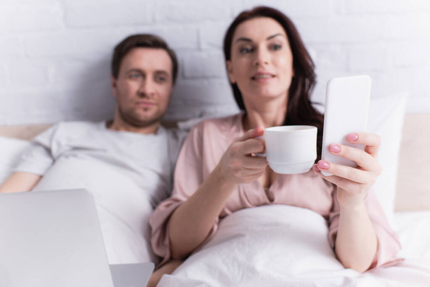 Smartphone in hand of woman with coffee lying near laptop and husband on bed  - Foto, immagini
