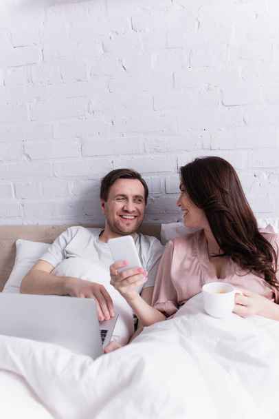 Smiling woman with cup showing smartphone to husband with laptop on bed  - Zdjęcie, obraz