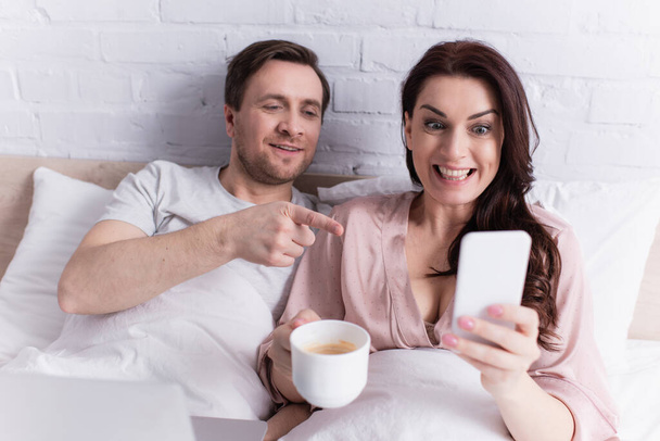 Cheerful woman with coffee holding smartphone near husband pointing with finger and laptop in bedroom  - Foto, immagini