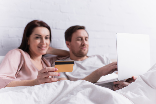 Hitelkártya a kezében egy nő fekszik közel férj laptoppal elmosódott háttér az ágyban  - Fotó, kép
