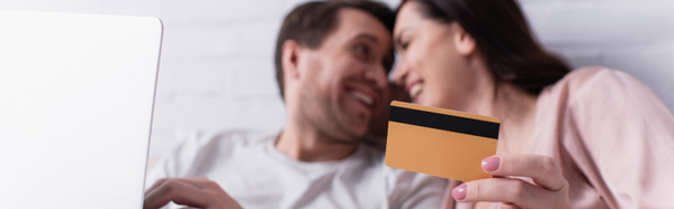 Credit card in hand of woman smiling near husband with laptop on blurred background, banner  - Photo, Image