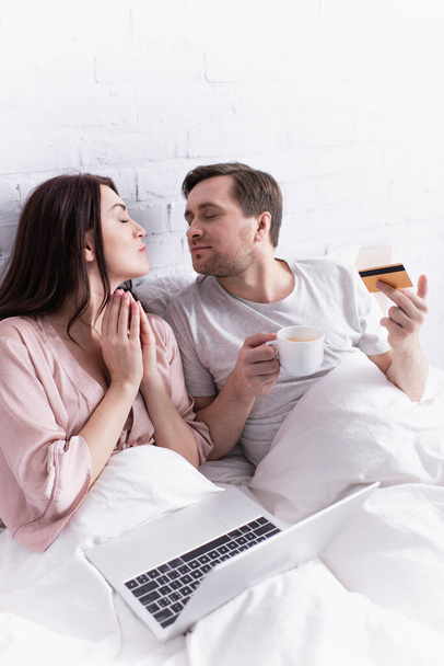 大人のカップルでクレジットカード、コーヒー、ノートパソコンがベッドの上に座って  - 写真・画像