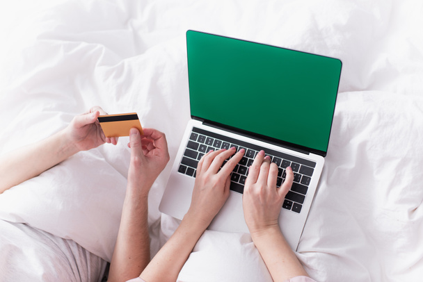 Vista ritagliata della donna che utilizza laptop con chiave cromatica vicino al marito con carta di credito sul letto  - Foto, immagini
