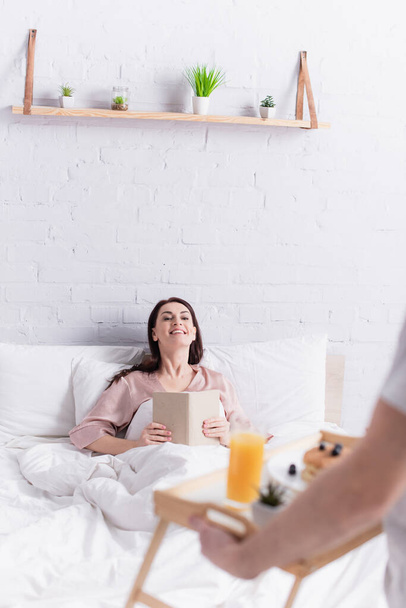 Mujer alegre sosteniendo libro en la cama cerca del marido con desayuno en bandeja en primer plano borrosa  - Foto, imagen
