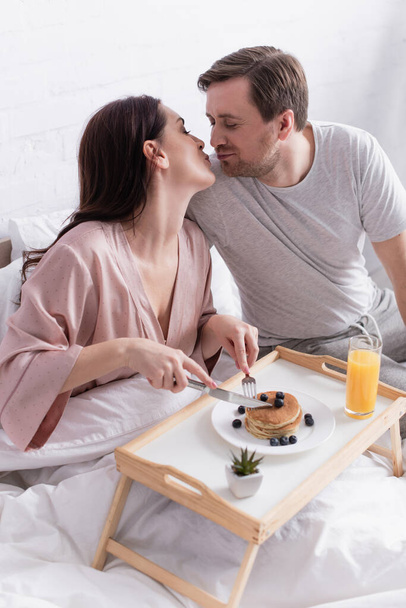 Woman with cutlery cutting pancakes and kissing husband in bedroom  - Foto, afbeelding