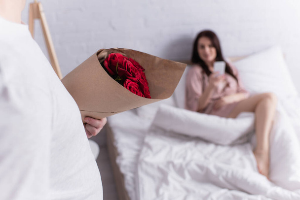 Man holding bouquet of roses near wife on bed on blurred background  - Valokuva, kuva