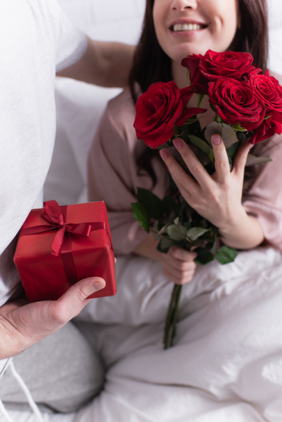 Обрізаний вид усміхненої жінки, що тримає троянди біля чоловіка з подарунком на ліжку
  - Фото, зображення