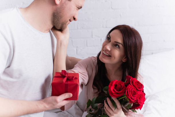 Smiling woman with roses touching face of husband with present on bed  - Фото, изображение