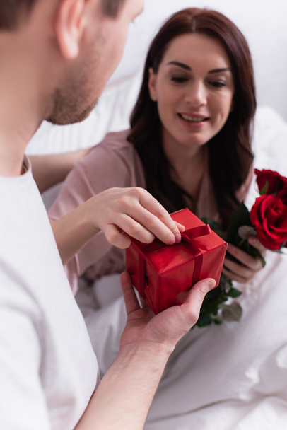 Man holding present near wife with roses blurred on bed  - Φωτογραφία, εικόνα