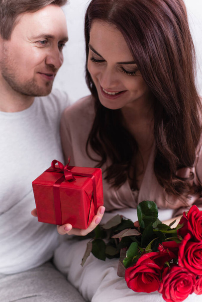 Smiling woman holding gift box and roses near husband in bedroom  - Фото, изображение