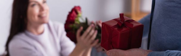 Man holding present and giving flowers to wife on blurred background, banner  - Foto, imagen