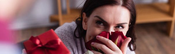 Brunette woman smelling roses near husband with present blurred on foreground, banner  - Fotoğraf, Görsel
