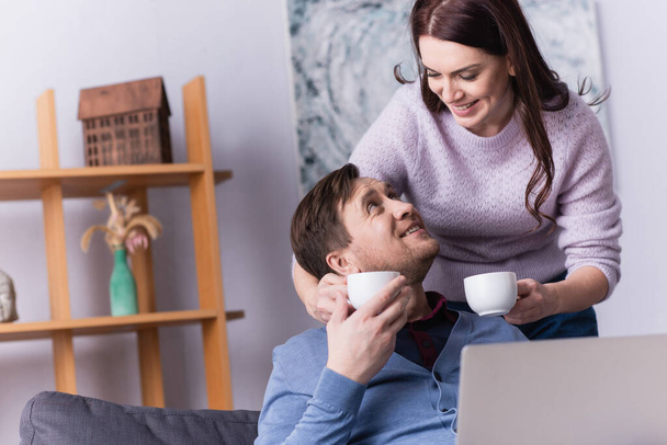 Woman holding cups of coffee near husband with laptop  - Foto, Imagem