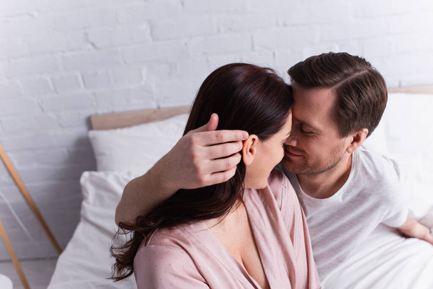 Dospělý muž dotýká vlasy brunetky manželky v pyžamu na posteli  - Fotografie, Obrázek