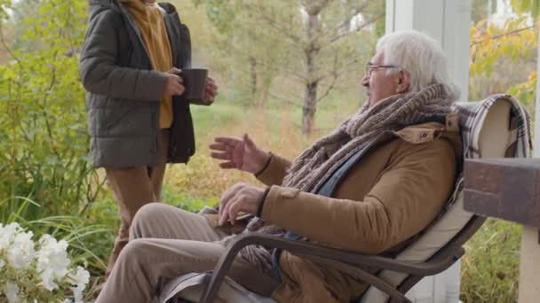 Foto media dell'anziano caucasico che indossa vestiti caldi e occhiali da vista seduto su una sedia a dondolo in terrazza. Poi suo nipote adolescente lo saliva una grande tazza di tè caldo - Filmati, video