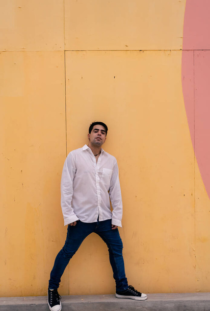 Full body photograph of a young Latino guy leaning against a plain colored wall. Urban clothing. - Photo, Image