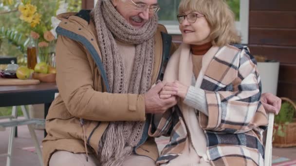 Inclinazione di felici anziani sposi caucasici indossando vestiti caldi seduti sulla terrazza della loro casa, tenendosi per mano e guardando la fotocamera - Filmati, video