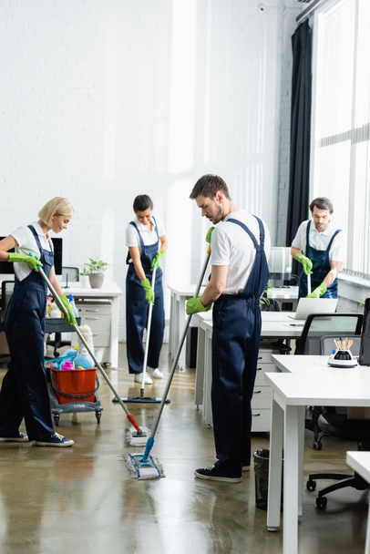 Interracial cleaners in overalls cleaning floor in office  - Foto, Bild