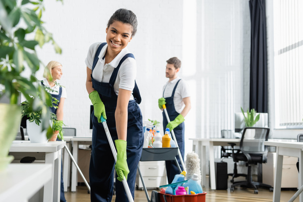 African american cleaner holding mop near colleagues and plant in office  - Photo, Image