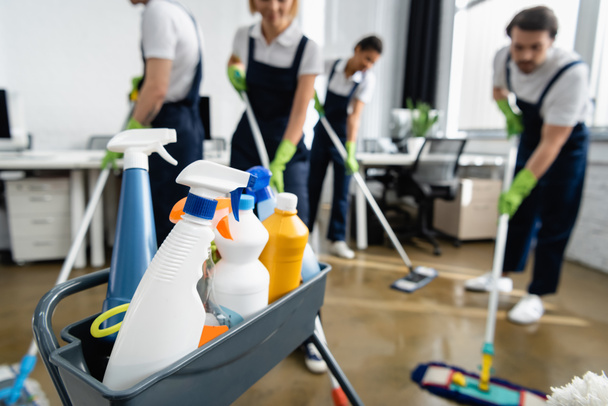 Garrafas com detergentes perto de limpadores trabalhando em fundo desfocado no escritório  - Foto, Imagem