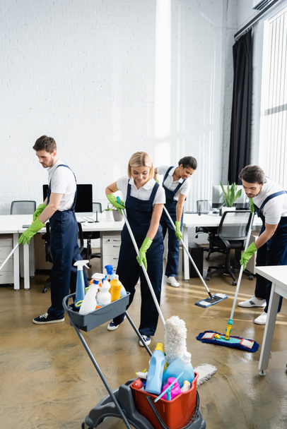 Gülümseyen ırklar arası temizleyiciler ofiste deterjanların yanında yerleri yıkıyorlar.  - Fotoğraf, Görsel