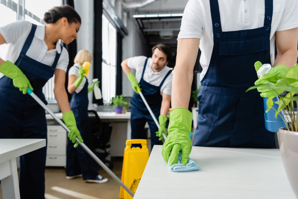 Cleaner cleaning table near plant and multiethnic colleagues on blurred background in office  - Photo, Image
