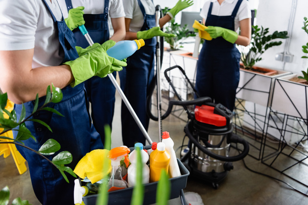 Cropped view of cleaner in rubber gloves holding detergent near colleagues and vacuum cleaner in office  - Foto, Imagem