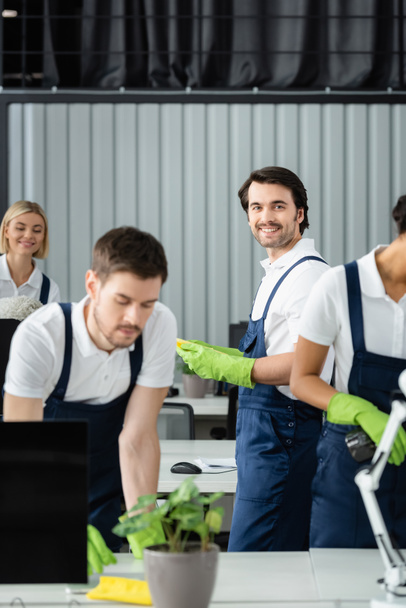 Smiling cleaner in rubber gloves looking at camera near interracial colleagues in office  - Photo, Image