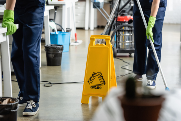 Oříznutý pohled na mokré podlahové tabule v blízkosti čističů pracující v kanceláři  - Fotografie, Obrázek